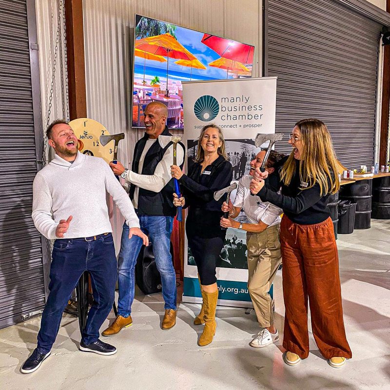 Group of people laughing and holding axes indoors near Manly Business Chamber banner.