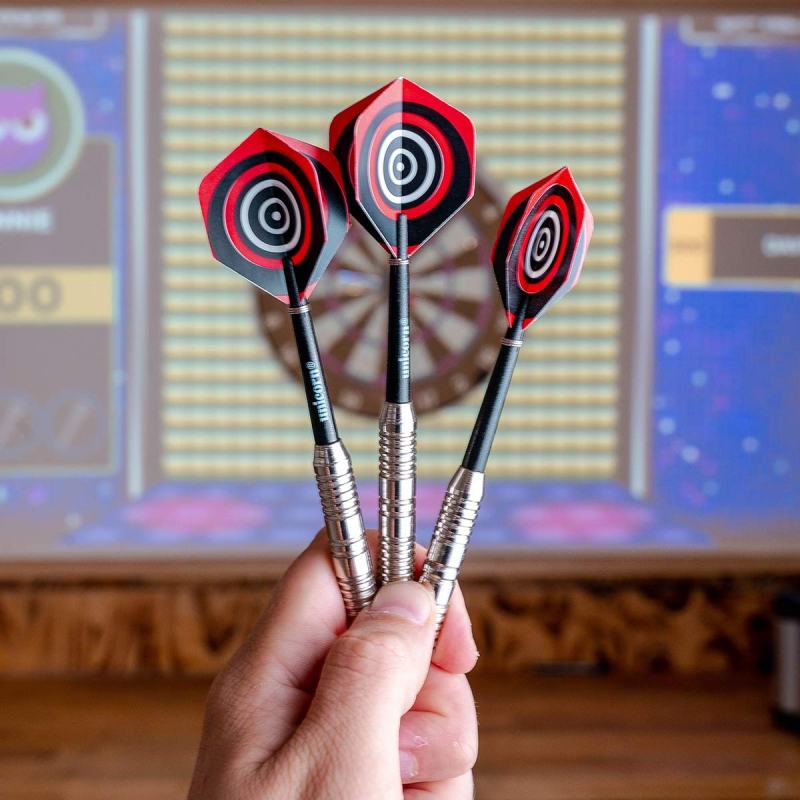 Hand holding three darts with red and black flights, dartboard in background.