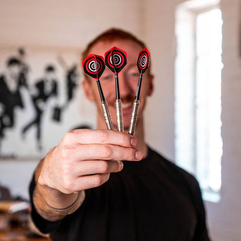Person holding three darts towards the camera in a room with a painting on the wall.
