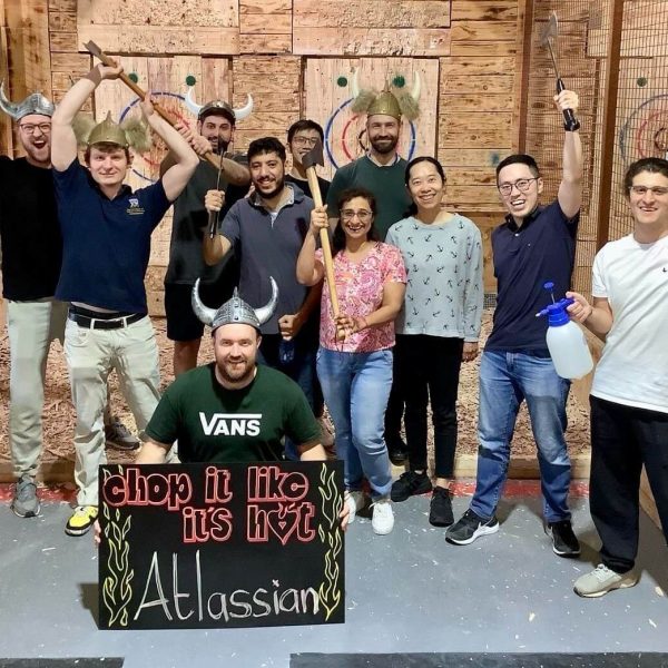 Group of people with axes at an axe-throwing venue, holding a 'chop it like it's hot' sign.