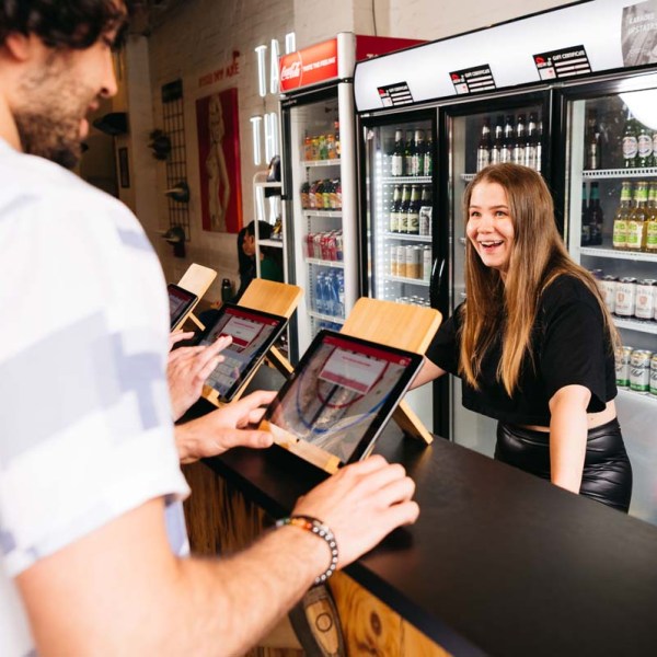 Woman smiling at a customer using a tablet at a store counter with beverage fridges in the background.