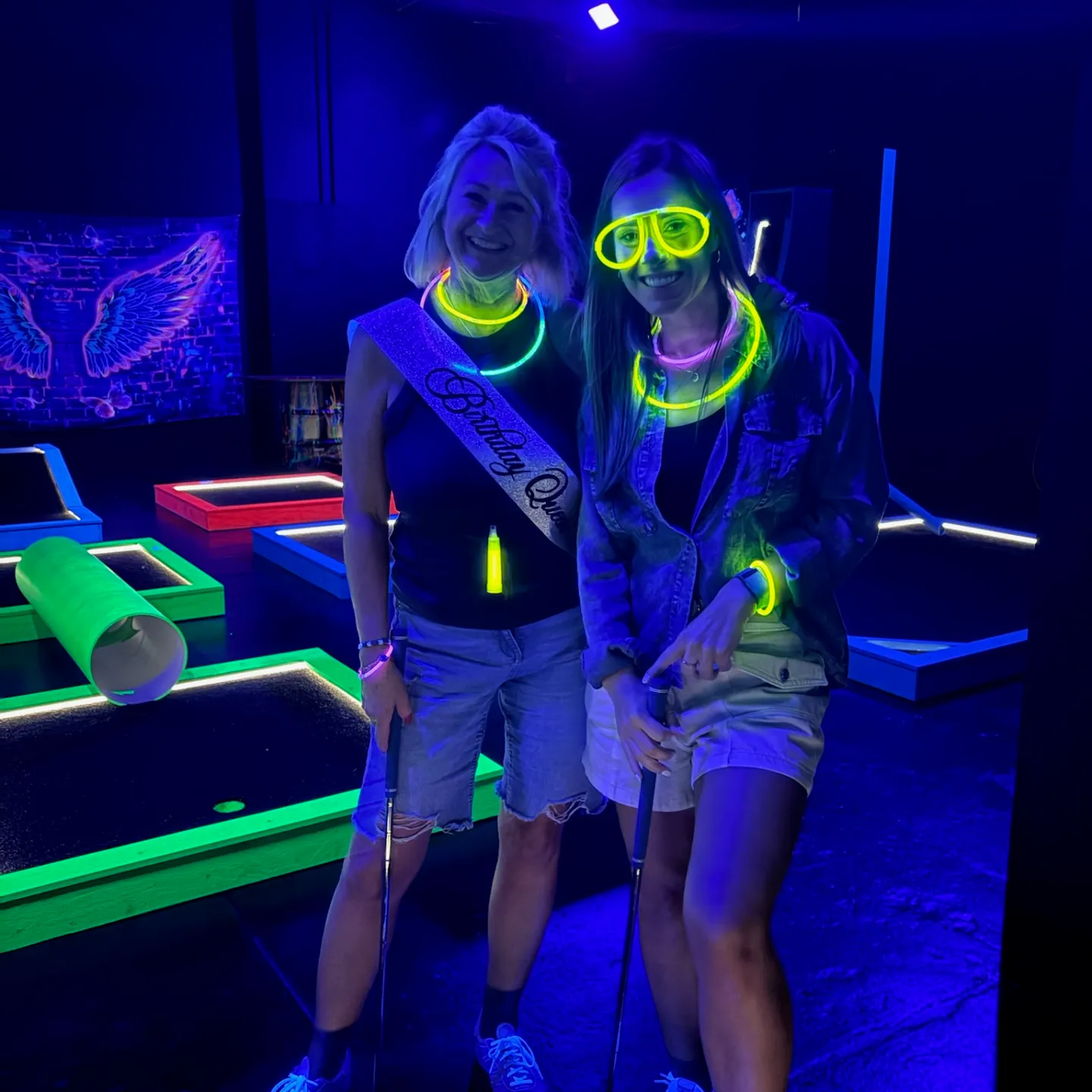 Birthday girl and friend playing glow in the dark mini golf with neon necklaces, bracelets and glasses