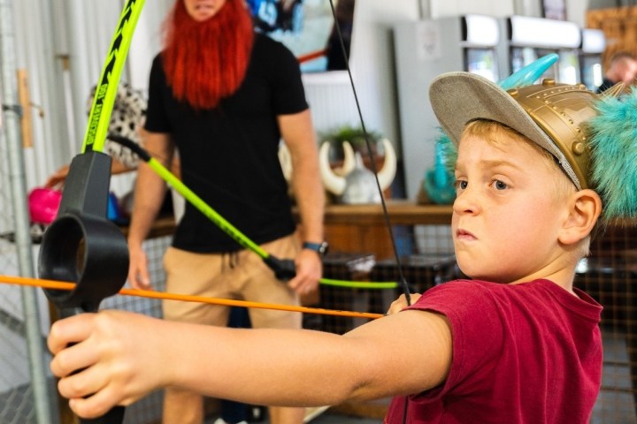 young boy aiming a bow and arrow inside at target practice