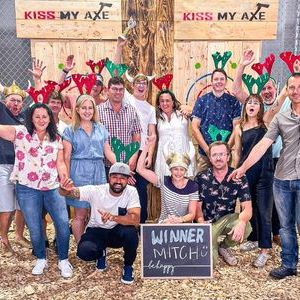 Group photo of 14 people wearing festive hats, holding a sign that says 'Winner Mitch' at an axe-throwing venue.