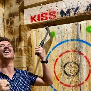 Man celebrating near target after axe throw with 'KISS MY AXE' sign above.