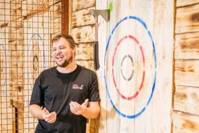 Man laughing near a wooden axe throwing target with a hatchet on it.