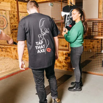 Man in 'Tap That Axe' shirt and woman with axe in indoor lumber setting.