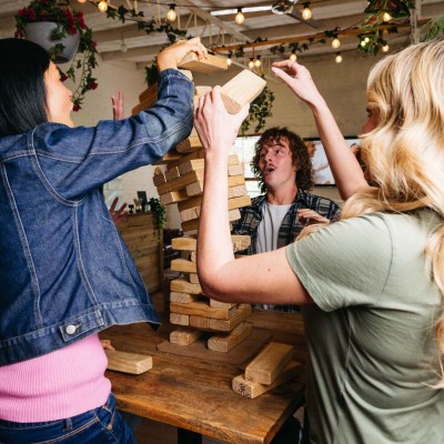 Three people playing a game of giant Jenga indoors, with plants and string lights overhead.