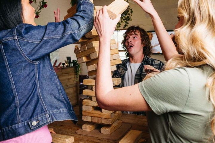 Three people playing a game of giant Jenga indoors, with plants and string lights overhead.