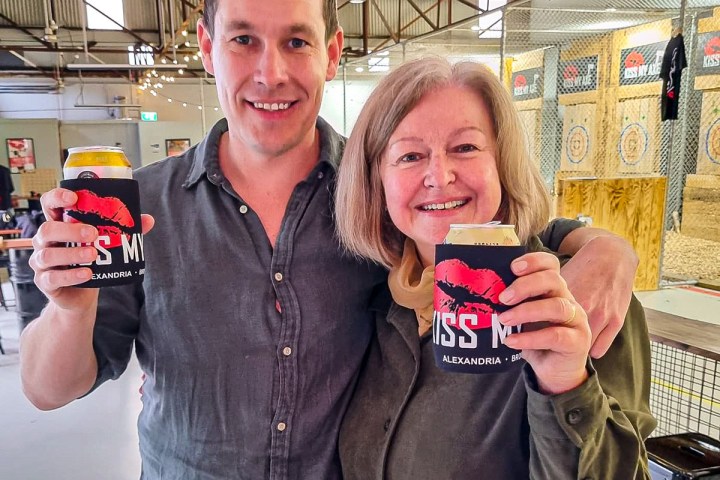 Two people smiling indoors, holding canned drinks with black and red koozies.