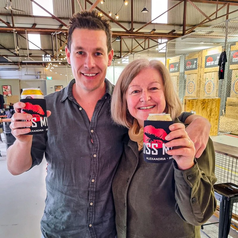 Two people smiling indoors, holding canned drinks with black and red koozies.
