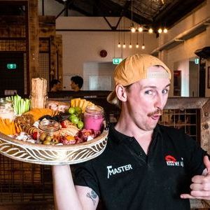 Person in cap holding a platter with colorful fruits and snacks indoors.