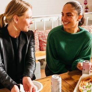 Two women sitting at a table eating pizza, smiling and talking.