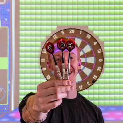 Person holding darts in front of face, with dartboard and colorful screen in the background.