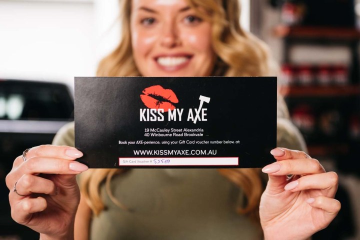 Person holds a 'Kiss My Axe' gift card, smiling in the background.
