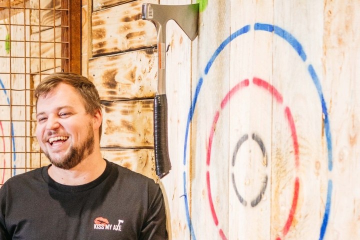 Man smiling near an axe embedded in a wooden target with colorful rings.
