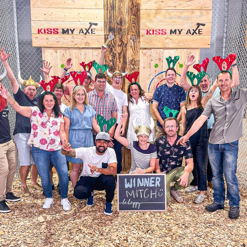 Group of people posing with antler hats and 'Kiss My Axe' sign indoors.