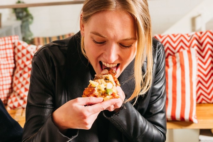 Person in black jacket eating a slice of pizza indoors with red chevron pillows in the background.