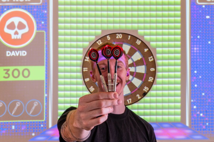 Person holding three darts in front of a dartboard with digital scoreboard.