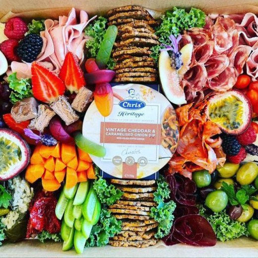 Colorful charcuterie board with meats, cheeses, fruits, veggies, and dip surrounded by greenery.
