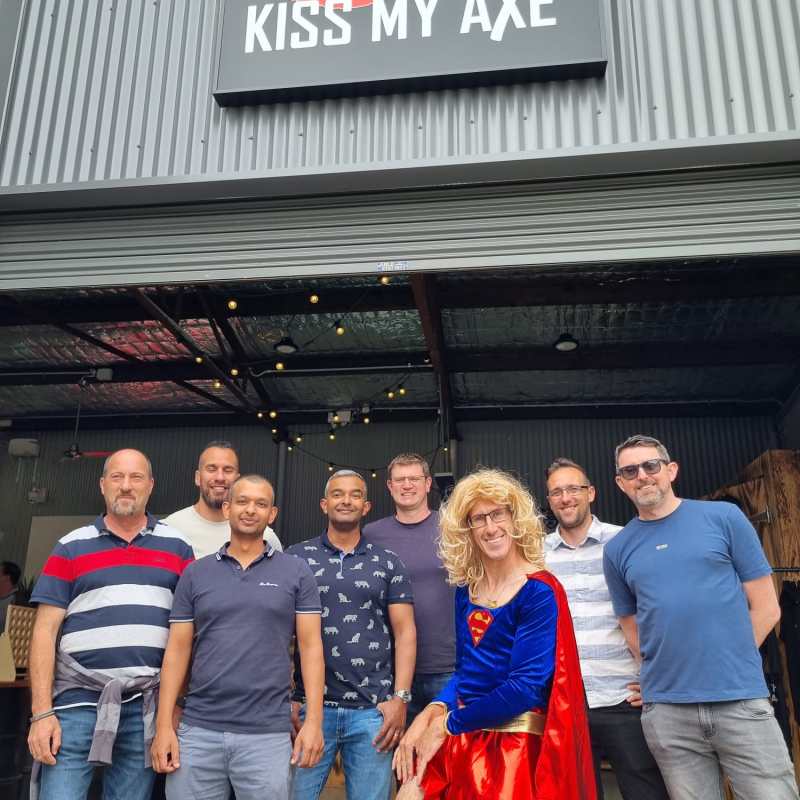 Group of eight people posing in front of 'Kiss My Axe' sign, one in a superhero costume.