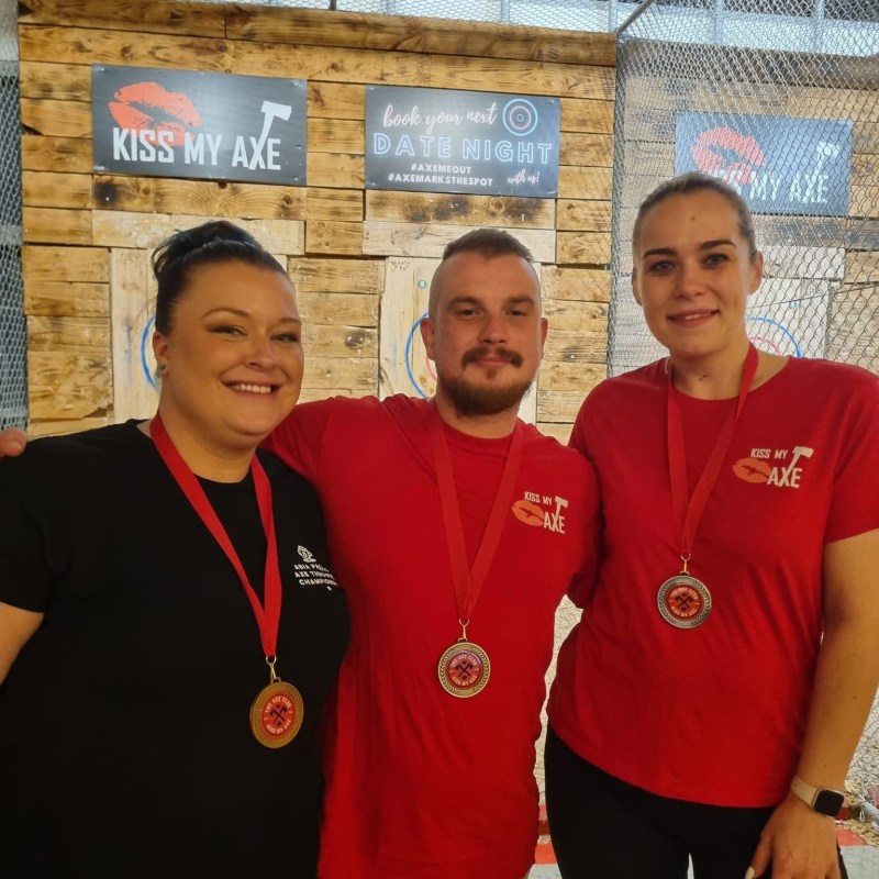 Three people with medals standing in front of an axe throwing area.
