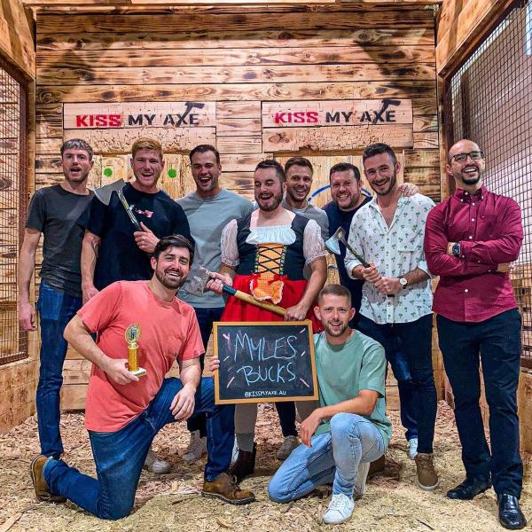 Group of men at axe throwing venue, one in costume, holding axes and a sign reading 'Myles Bucks'.
