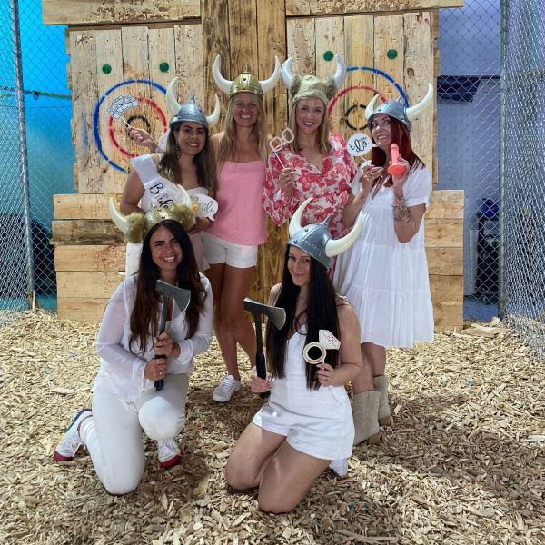 Six women wearing Viking helmets posing with axes in front of wooden targets, standing on wood chips.