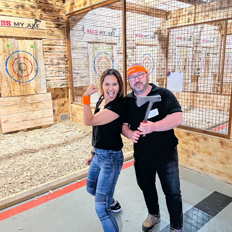 Two people at an axe throwing venue, one flexing arm and the other holding an axe.