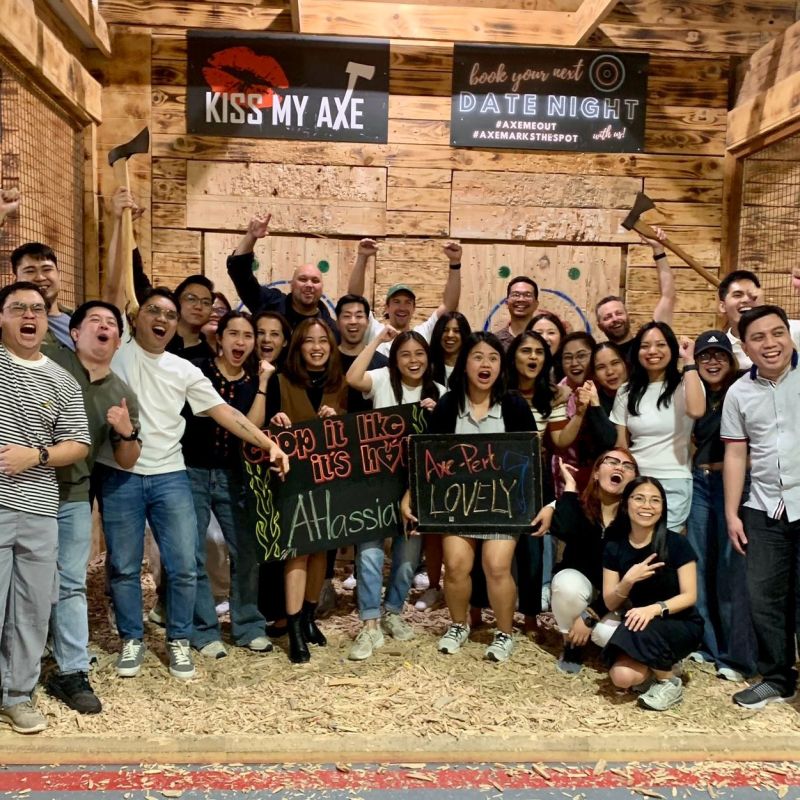 Group of people cheering at axe throwing venue, holding signs, with wooden targets in the background.