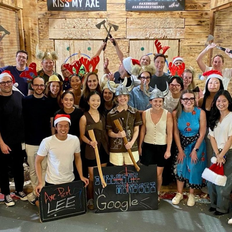 Group of people posing with axes, wearing hats and festive attire, in an indoor axe-throwing venue.