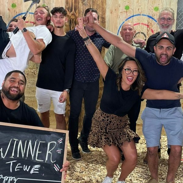 Group of people celebrating at an axe-throwing venue, one holding a winner sign, with wood targets behind.