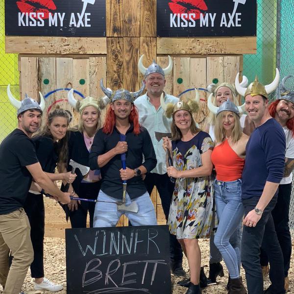 Group with axe, viking hats, smiles, beside sign 'Winner Brett' at axe-throwing venue with logo 'Kiss My Axe'.