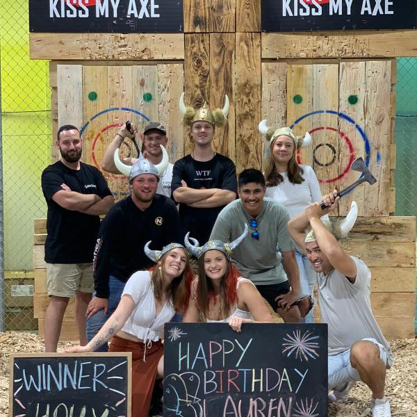 Group celebrating with axes, wearing horned hats, holding signs, in front of target boards.
