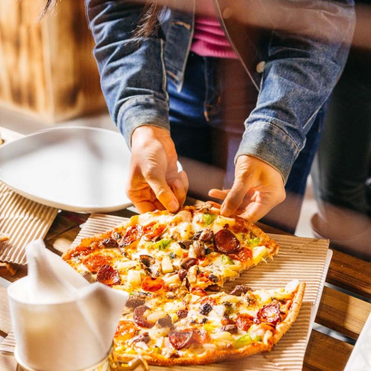 Person in denim jacket taking slice from a pizza on a wooden table.