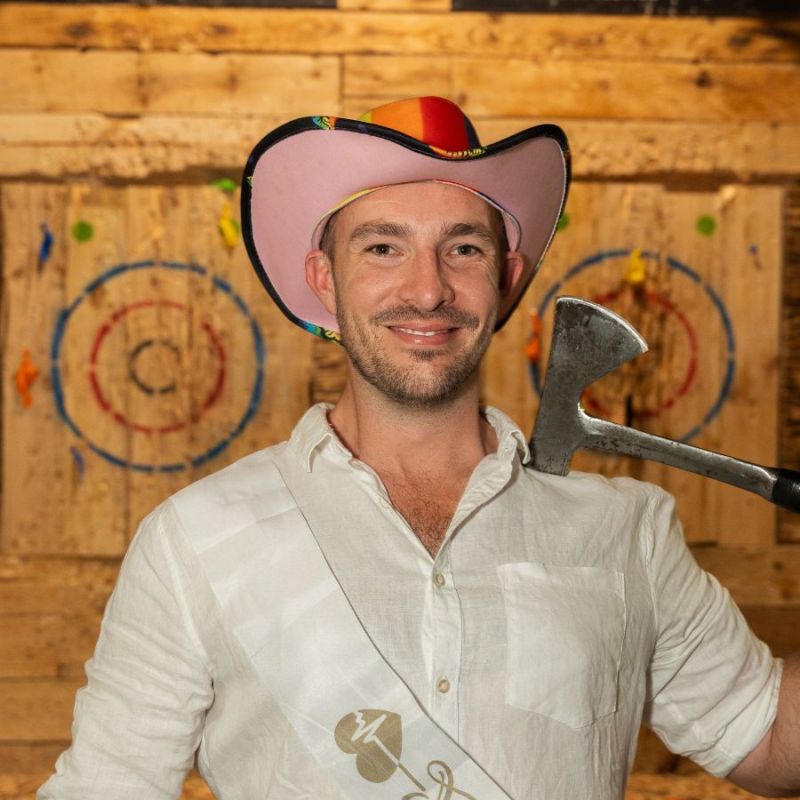 Man with a pink cowboy hat holding an axe in an axe-throwing venue.