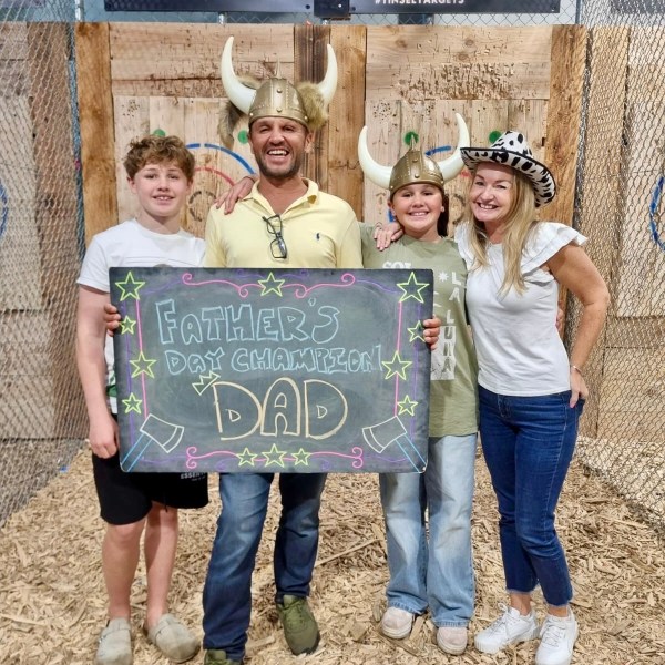Four people smiling, holding a Father's Day sign with axe-throwing targets in the background.