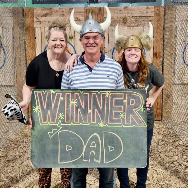 Three people in helmets, one holds a 'Winner Dad' sign at an axe-throwing venue.