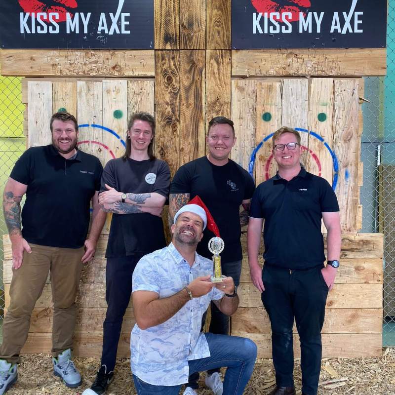 Five people posing by wooden target boards, one kneeling with a trophy, in a venue with 'Kiss My Axe' signs.