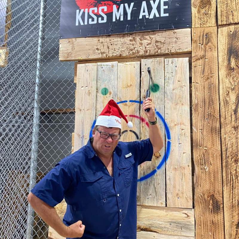 Man in Santa hat posing with axe near 'Kiss My Axe' sign.