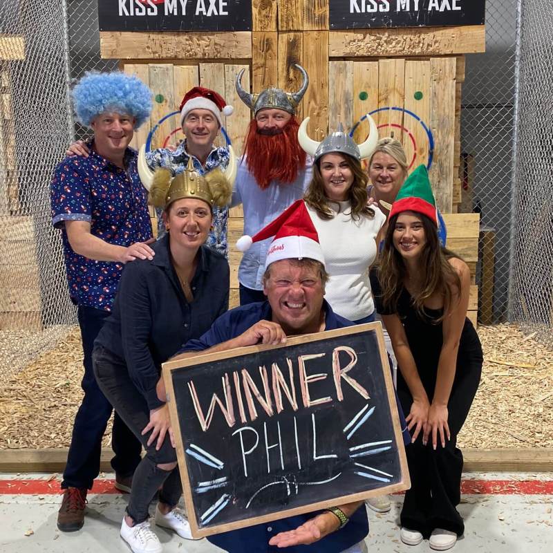 Group of people in festive hats holding 'Winner Phil' sign at axe-throwing venue.