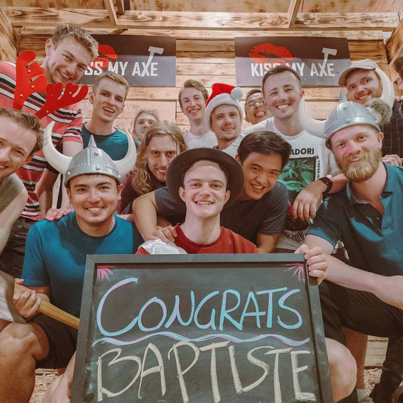 Group of 14 people at an axe-throwing event wearing hats, holding a 'Congrats Baptiste' sign.