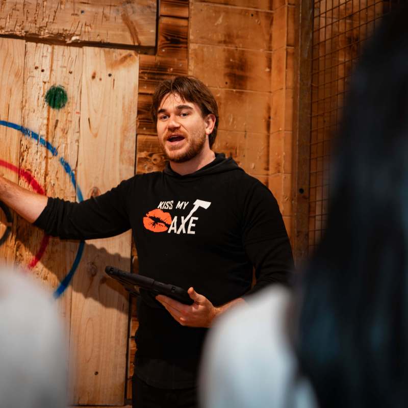 Man explaining axe throwing at a wooden target with people listening.