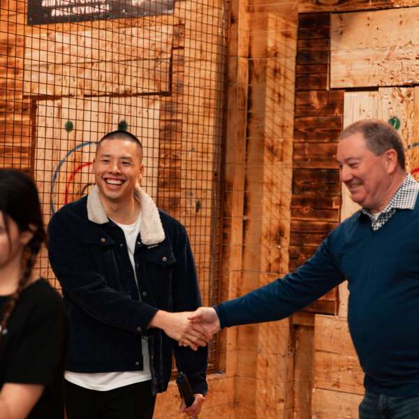 Two men shake hands at an axe-throwing venue, with a woman walking past.