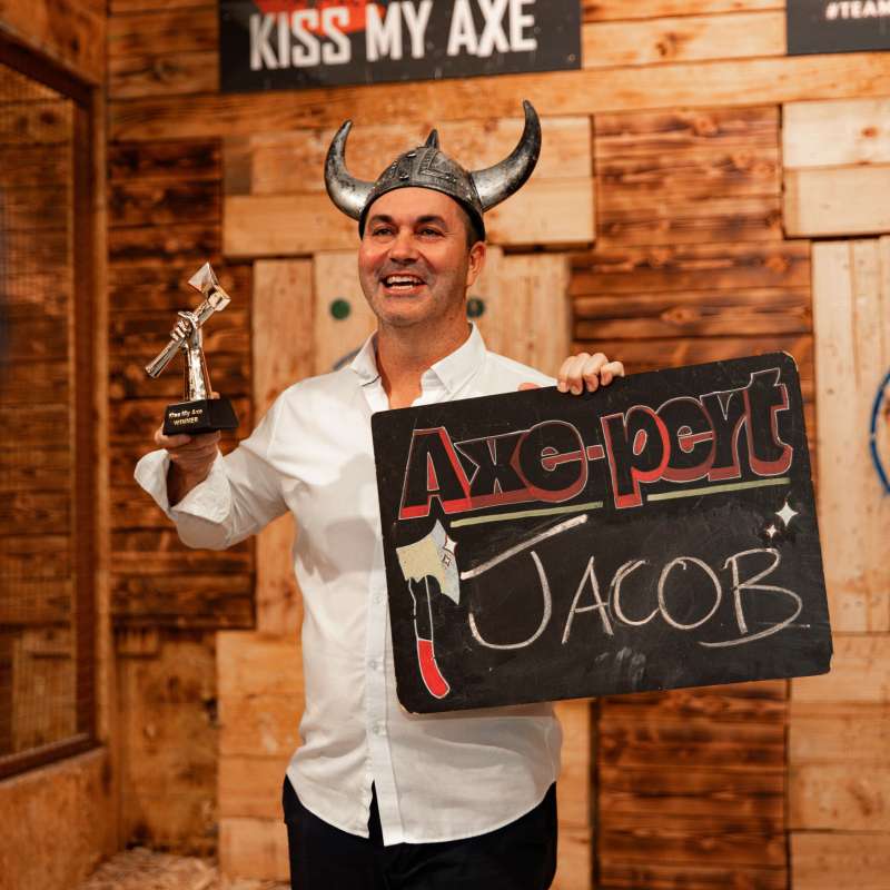 Smiling man in helmet holds trophy and 'Axe-pert Jacob' sign in axe-throwing venue.