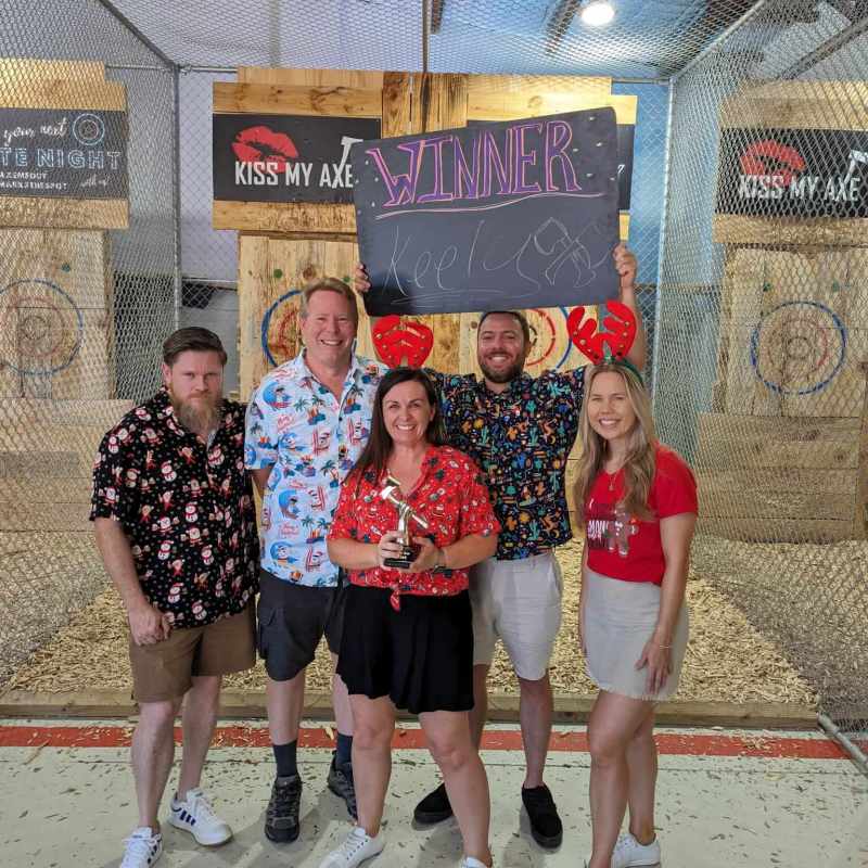Group celebrating at an axe-throwing venue, person holding 'Winner' sign.