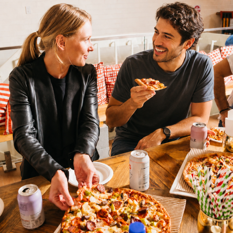Two people eating pizza at a wooden table with drinks and pizza boxes.