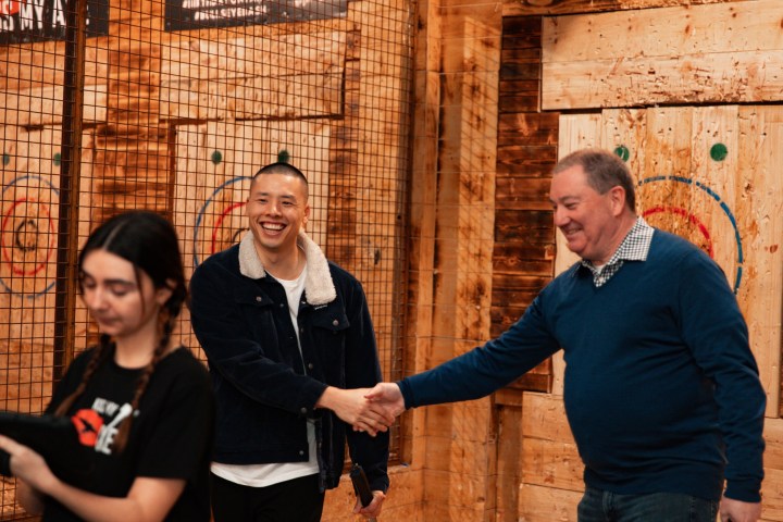 Two men shake hands, smiling, in a wood-paneled room.