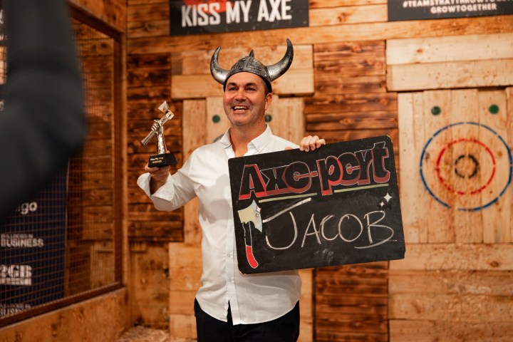 Man in Viking helmet holds trophy and 'Axepert Jacob' sign in axe-throwing venue.