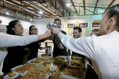 Group of people clinking glasses over pizza boxes, smiling in a casual setting.